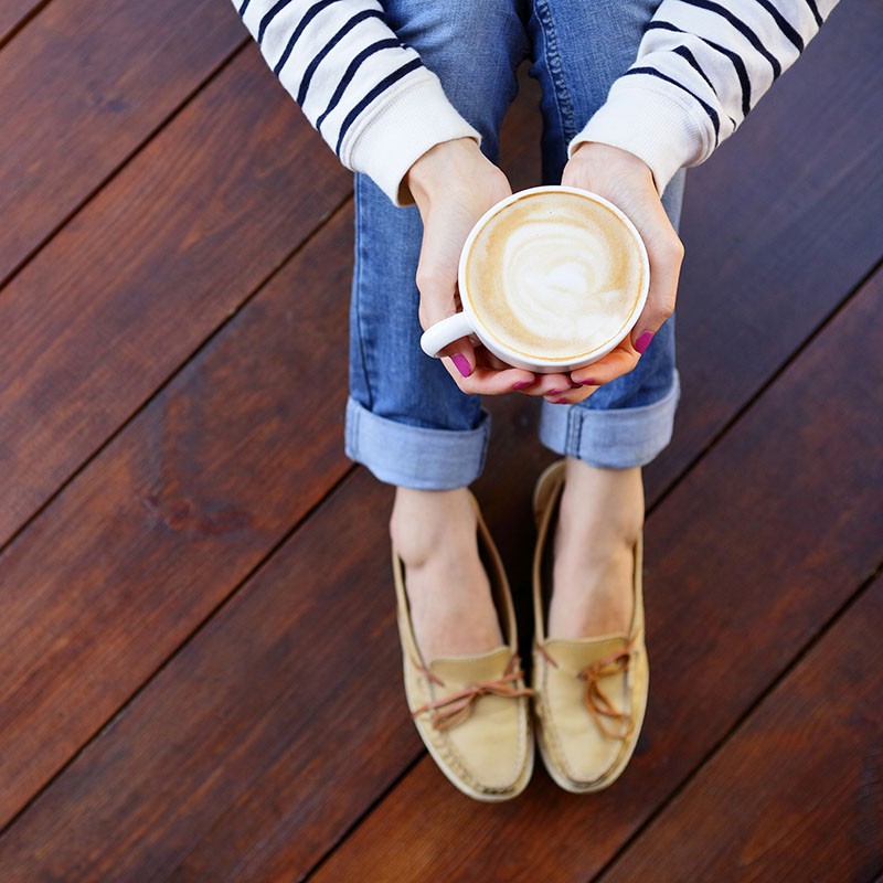 Woman holding in hands cup of coffee with milk sitting on the wooden floor, top view point