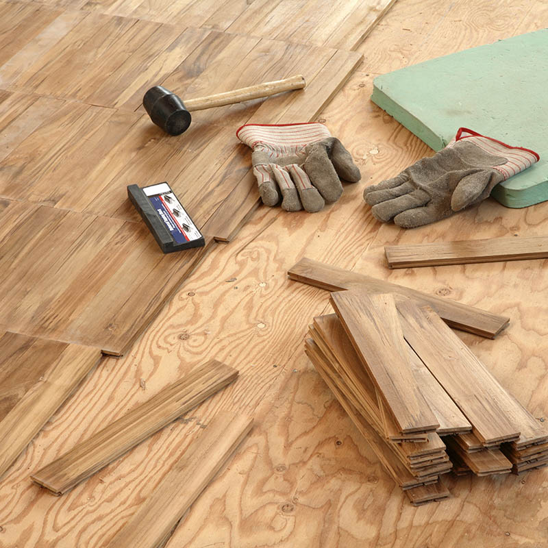 Laying wood flooring.  Newly laid flooring stretches in a diagonal line across left half of frame .  Subfloor on right with a pair of work gloves, a rubber hammer and a black plastic tapping block spread before a green kneeing mat.  A stack of wood to be laid in front and small scattering of individual pieces around it on subfloor which fills right of frame.